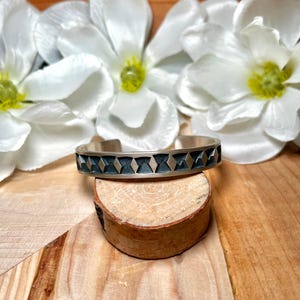 May include: A silver cuff bracelet with a geometric pattern of dark blue and silver diamonds. The bracelet is displayed on a small wooden slice, with white flowers in the background.