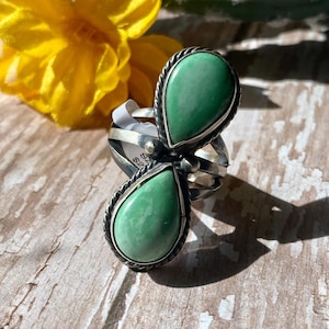 May include: A silver ring featuring two teardrop-shaped green stones set in twisted metal borders. The ring has a split band design with a polished finish, showcasing a dark patina in the crevices of the metalwork surrounding the smooth stones.