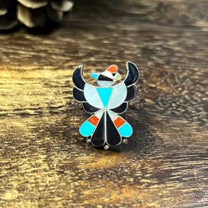 May include: A silver ring featuring a turquoise, red, white and black inlay design of a stylised bird. The ring is set against a wooden background.