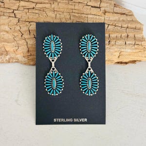 May include: A pair of sterling silver earrings with a turquoise stone design. Each earring features an oval top and a teardrop-shaped bottom, both adorned with small turquoise stones. The earrings are displayed on a black card with the text "STERLING SILVER".