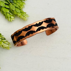 May include: A copper cuff bracelet with a textured surface and a black, patterned design. The open-ended bracelet is a statement piece, perfect for adding a touch of artistry to any outfit. The bracelet is approximately 1 inch wide.