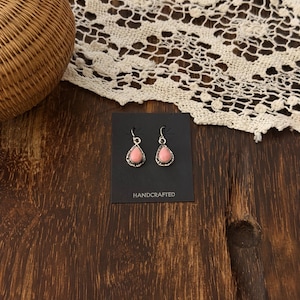 May include: A pair of silver-toned drop earrings featuring teardrop-shaped pink stones set in textured metal frames. The earrings are attached to a black rectangular display card with the word HANDCRAFTED printed in white at the bottom center.