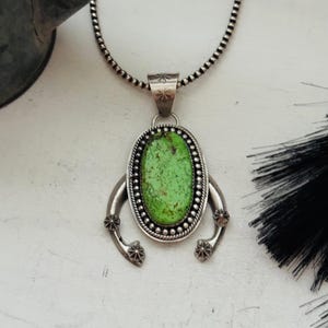 May include: A sterling silver necklace featuring an oval-shaped, vibrant lime green stone pendant. The pendant is framed by a beaded border and accented with two curved, star-stamped silver arms. It hangs from a matching silver ball chain.