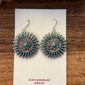 May include: A pair of Zuni handmade silver earrings. Each earring features a central turquoise stone surrounded by a silver design with turquoise accents. The earrings are on a white card with the text "ZUNI HANDMADE JEWELRY".