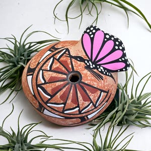 May include: A terracotta-colored, round decorative item with a painted geometric design in red, white, and black. A pink and black butterfly is attached to the top. Green air plants surround the item, creating a natural, artistic display.
