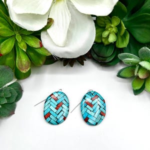 May include: A pair of oval-shaped dangle earrings featuring a woven herringbone pattern in shades of turquoise blue and deep red. The earrings are suspended from thin silver-toned metal hooks and display a smooth, polished finish on the surface.