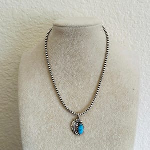 May include: A silver necklace with a beaded chain and a turquoise pendant. The pendant is oval-shaped and set in a silver frame with a leaf design. The necklace is displayed on a mannequin.