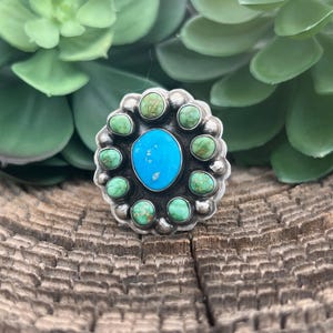 May include: A silver ring with a central turquoise stone surrounded by smaller green turquoise stones. The ring has a floral design and is set against a wooden background with green foliage.
