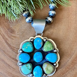 May include: A silver pendant necklace with a large, flower-shaped turquoise and green stone pendant. The pendant is surrounded by a silver frame and is strung on a beaded silver necklace. The necklace is displayed on a wooden surface.