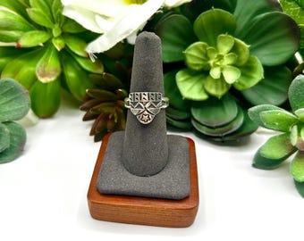Könnte beinhalten: Ein silberner Ring mit einer schildförmigen Vorderseite, die ein eingraviertes geometrisches Muster und einen kleinen Stern in der Mitte aufweist. Der Ring steht auf einem grauen, kegelförmigen Ständer mit Holzsockel vor grünen Sukkulenten.