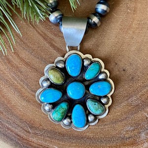 May include: A silver pendant necklace with a flower design. The pendant features turquoise and green stones set in a scalloped silver frame. The necklace has a beaded silver chain. The background is a wooden surface.