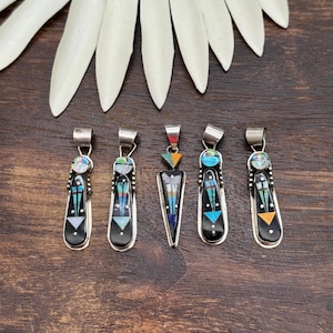 May include: Five silver pendants featuring inlaid stone designs. Each pendant displays a black base with geometric patterns in blue, orange, and white. The pieces include circular and triangular shapes with silver bails at the top for hanging.