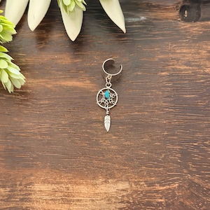 May include: A silver ear cuff featuring a dreamcatcher design with a small turquoise stone in the center and a dangling feather charm. The jewelry is set against a dark wood grain background with green foliage.