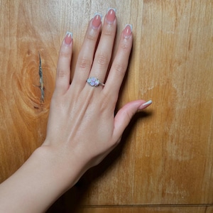 May include: A silver ring with a cluster of small, light-colored gemstones is worn on a hand with manicured nails. The ring's design features a floral arrangement of the gemstones. The nails are painted with a light pink and white color.