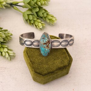 Puede incluir: Un brazalete de plata con una piedra turquesa ovalada central con patrones de matriz marrón. La banda de metal muestra un diseño repetitivo de óvalos y puntos estampados a los lados, con un acabado pulido y detalles oscurecidos en relieve.