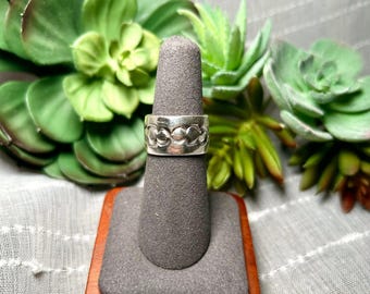 Könnte beinhalten: Ein silberner Bandring mit einem sich wiederholenden Muster aus geprägten kreisförmigen Formen auf der Außenseite. Der Ring wird auf einem grauen, kegelförmigen Ständer auf einem quadratischen Holzsockel präsentiert und zeigt eine metallische Oberfläche.