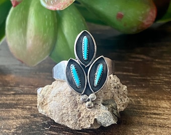 May include: A silver ring with three turquoise stones set in a leaf design. The ring is sitting on a small rock.