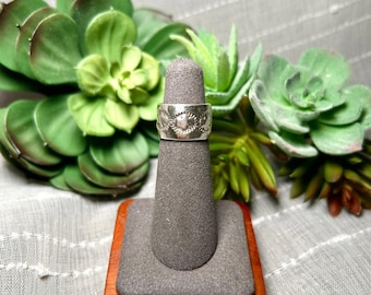 Könnte beinhalten: Ein silberner Bandring mit einem sich wiederholenden geprägten Sonnen- oder Blumenmuster auf der Außenfläche. Der Ring wird auf einem grauen, kegelförmigen Ständer mit quadratischem Holzsockel präsentiert und aufrecht positioniert.