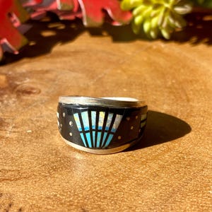 May include: A silver ring with a black band featuring a geometric design with turquoise and white accents. The ring has a unique, artistic style, with small dot details. The ring is set on a wooden surface.