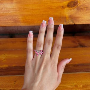 May include: A hand wearing a silver wire ring with small pink beads. The ring has a spiral design. The nails are painted with a French manicure. The background is a wooden surface.