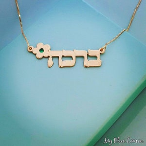 May include: Gold necklace with a flower charm and the Hebrew word "Sarah" in cursive lettering. The flower charm has a green gemstone in the center.