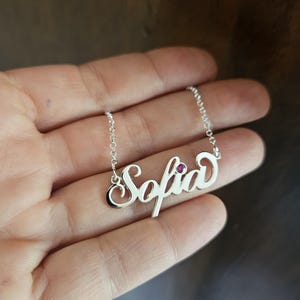 May include: A silver-toned name necklace featuring the name Sofia in a cursive script font. The necklace includes a small, round, dark pink gemstone accent embedded within the letter i. The pendant is attached to a delicate silver-toned chain.