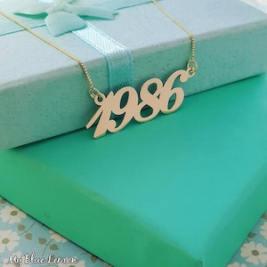Peut inclure: Collier en or avec un pendentif 1986 en cursive sur un fond bleu turquoise.