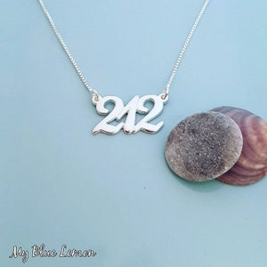 Könnte beinhalten: Eine silberne Halskette mit der Zahl "212" in Schreibschrift. Die Halskette hängt an einer silbernen Kette. Der Hintergrund ist hellblau, mit zwei grauen Steinen und einer Muschel.