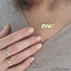 Könnte beinhalten: Eine goldene Halskette mit einem Anhänger, auf dem "1986" in einer kursivschriftlichen Schriftart steht.
