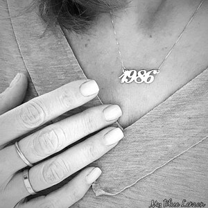 Könnte beinhalten: Eine silberne Halskette mit einem Anhänger, auf dem "1986" in einer kursivschriftlichen Schriftart steht.