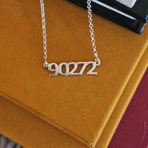 May include: A silver-colored necklace with the number "90272" as a pendant. The necklace is resting on a stack of books with a mustard-yellow cover. The chain is delicate and the numbers are in a sans-serif font.