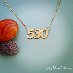 May include: Gold necklace with a pendant featuring the number 530. The necklace is on a light blue background.