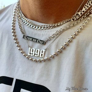 May include: A silver chain necklace with four layers. The top layer is a thick curb chain. The second layer is a delicate chain with a silver pendant that says "Trust no one". The third layer is a delicate chain with a silver pendant that says "1998". The bottom layer is a silver ball chain.