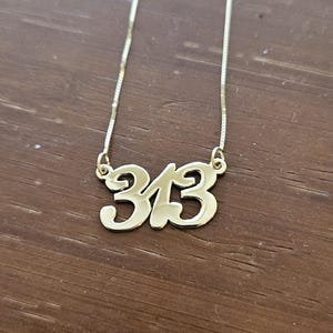 Puede incluir: Un collar dorado con el número "313" en fuente cursiva. El colgante cuelga de una cadena fina. El collar se muestra sobre una superficie de madera marrón.