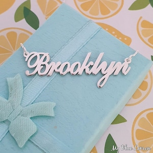 May include: Silver necklace with the name "Brooklyn" spelled out in a cursive font. The necklace is displayed on a light blue gift box with a light blue ribbon.