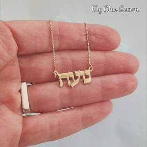 May include: A gold-coloured necklace featuring a pendant with Hebrew script lettering. The pendant is attached to a delicate box chain. The text My Blue Lemon is visible in the upper right corner of the image, which displays the jewellery piece.