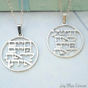 May include: Two silver pendant necklaces featuring Hebrew script within circular frames. The pendants are suspended from delicate silver chains. The necklaces are presented against a light backdrop, highlighting the detailed design and craftsmanship of the jewellery.
