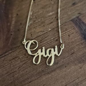 Può includere: Una collana color oro con un ciondolo a forma di nome in corsivo che riporta la scritta Gigi. Il ciondolo è fissato a una sottile catenina a maglia veneziana, con le lettere collegate tra loro in uno stile fluido e continuo in metallo.