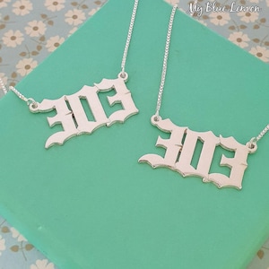 Könnte beinhalten: Zwei silberne Halsketten mit dem Wort "EIE" in gotischer Schrift. Die Anhänger hängen an feinen Silberketten, präsentiert auf einer türkisfarbenen Oberfläche. Der Hintergrund hat ein Blumenmuster und den Text "My Blue Lemon".