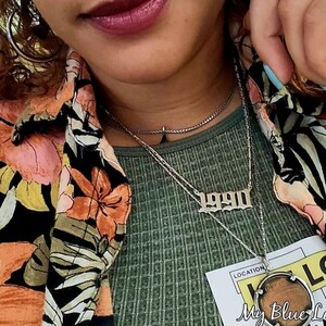 Könnte beinhalten: Eine Person trägt ein grünes Shirt mit einem floralen Hemd darüber. Sie trägt eine silberne Halskette mit einem 1990er-Anhänger und eine weitere silberne Halskette mit einem runden Anhänger.