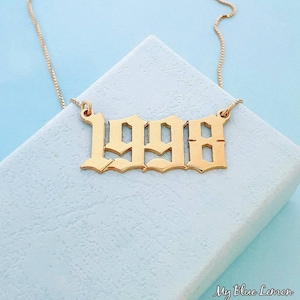 May include: Gold-colored necklace with the number "1998" in a gothic font. The pendant is suspended from a delicate chain. The necklace is displayed on a light blue surface.