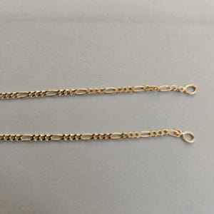 May include: Two parallel gold-toned metal chains featuring a classic Figaro link pattern. Each chain consists of a repeating sequence of three small circular links followed by one elongated oval link, terminating in a small circular jump ring at the end.