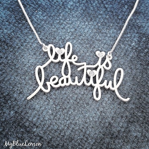May include: A silver necklace with the words "life is beautiful" in cursive script. The necklace has small heart-shaped accents and tiny gemstones. The chain is delicate and the background is a textured blue.