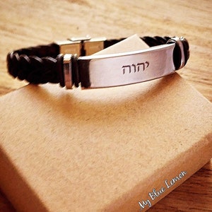 Pode incluir: Um bracelete de couro preto com uma placa de prata gravada com a palavra hebraica "מהה".