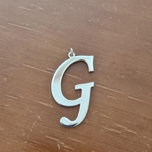 May include: A shiny silver pendant in the shape of the letter "G". The pendant has a loop at the top for a chain. The letter is in a cursive font and is displayed on a wooden surface.
