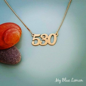 May include: Gold necklace with a pendant featuring the number 530. The necklace is on a light blue background.
