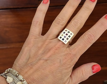 May include: A silver ring with a rectangular face featuring a grid pattern of small squares, each containing a different colored gemstone. The ring is on a hand with red nail polish, and a silver bracelet is visible on the wrist.