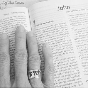 May include: A silver-toned metal ring featuring black Hebrew lettering, worn on a finger resting against an open page of a book titled John. The text on the page includes the heading The Word Becomes Flesh and the phrase Tree of Life Version.