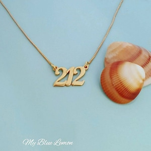 May include: A gold necklace with a pendant that says "212" on a light blue background. The necklace is next to two seashells.