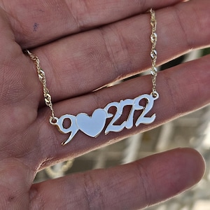 May include: A gold-colored necklace featuring a polished metal pendant with the number 9, a heart shape, and the number 272 in a connected script font. The pendant is attached to a thin, twisted gold-colored chain with a reflective metallic finish.